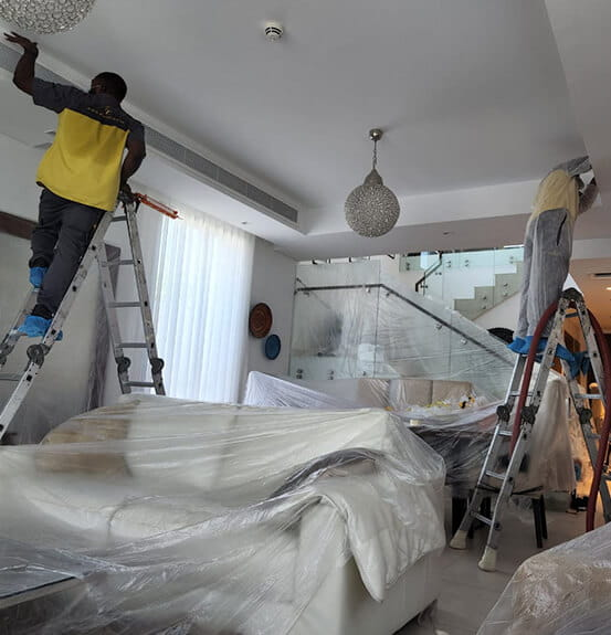 123 Two workers on ladders inspect ac ducts in a modern living room with furniture wrapped in plastic covers. The scene is busy and organised.