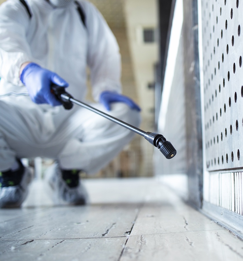 A person in protective gear and blue gloves crouches, spraying chemicals from a nozzle onto a perforated metal surface for pest control, conveying a focused, sterile environment.
