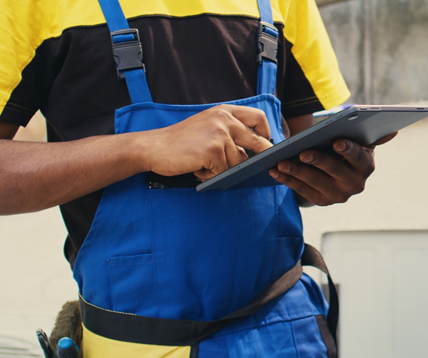 A person in blue and yellow overalls holds a tablet, standing near equipment outdoors. The focus is on their hands and attire, suggesting a technical task.