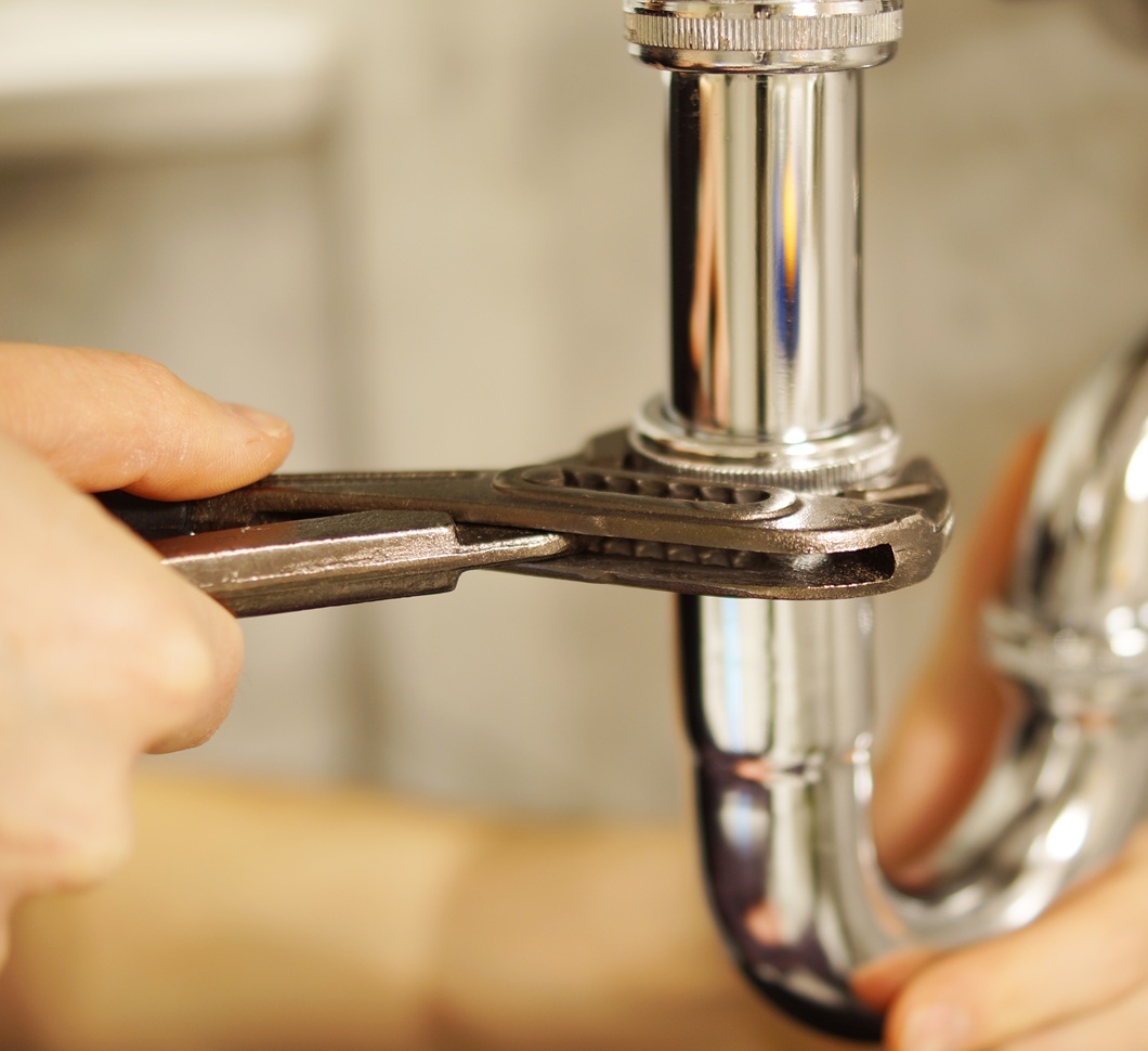 Hands tighten a shiny chrome pipe with a wrench, indicating a plumbing repair. The focus is on the tool and pipe against a blurred background.
