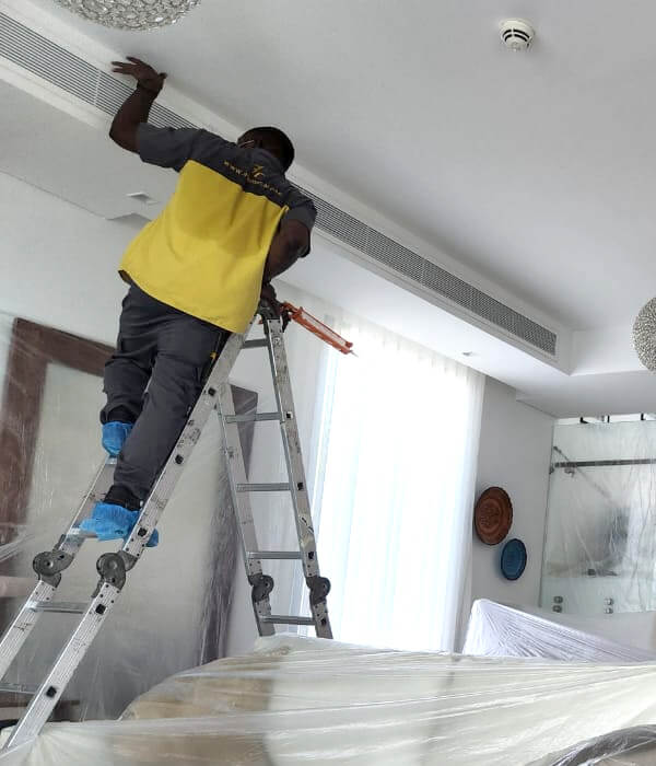 A worker stands on a ladder, wearing blue shoe covers and using a tool to fix a ceiling vent. The room is covered in protective sheets, conveying care and professionalism.