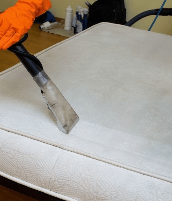 A person wearing an orange glove is using a vacuum cleaner attachment to clean a white mattress.