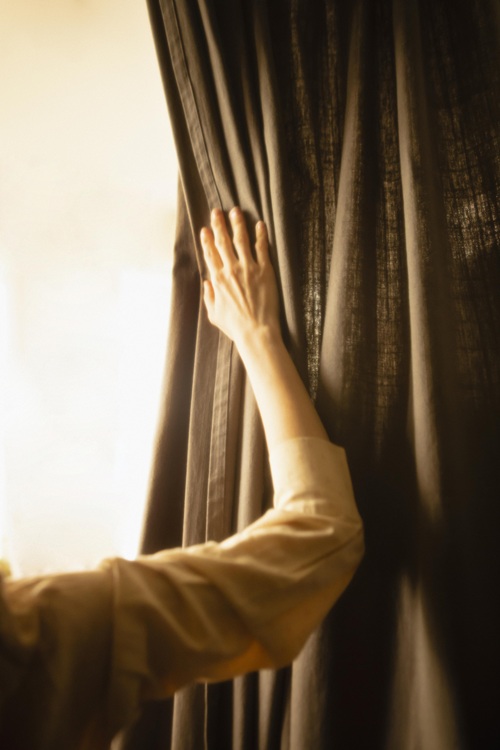 123 A person's hand gently reaches out to shape and restore the curtain’s natural drape on a dark curtain, backlit by warm, soft light, creating a calm and contemplative atmosphere.