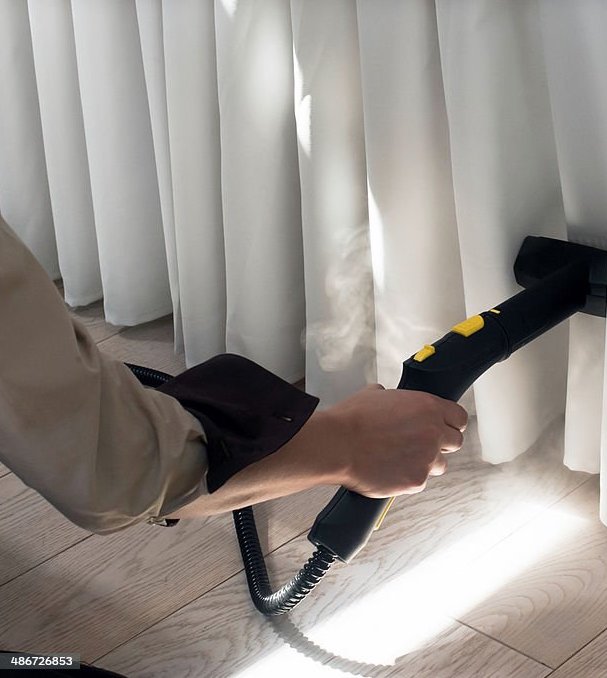 A person uses a steam cleaner on white curtains, with sunlight streaming through. The scene conveys cleanliness and focus, highlighting the cleaning process.