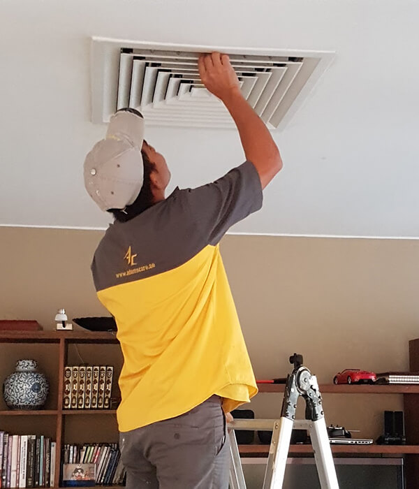 A person in a yellow and gray uniform from Adams Care stands on a ladder adjusting a ceiling vent. The background shows bookshelves with books and decorative items, such as a blue vase and a model car. The setting is calm and tidy.