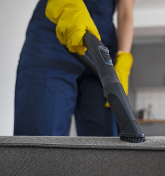 A person in blue overalls and yellow gloves uses a vacuum on a gray couch in a modern kitchen as part of furniture cleaning services. The setting conveys cleanliness and domestic focus.