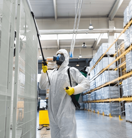 123 A person in a protective suit and gas mask sprays a white panel in a spacious warehouse. Shelves with boxes line the walls, conveying a clean-up task.