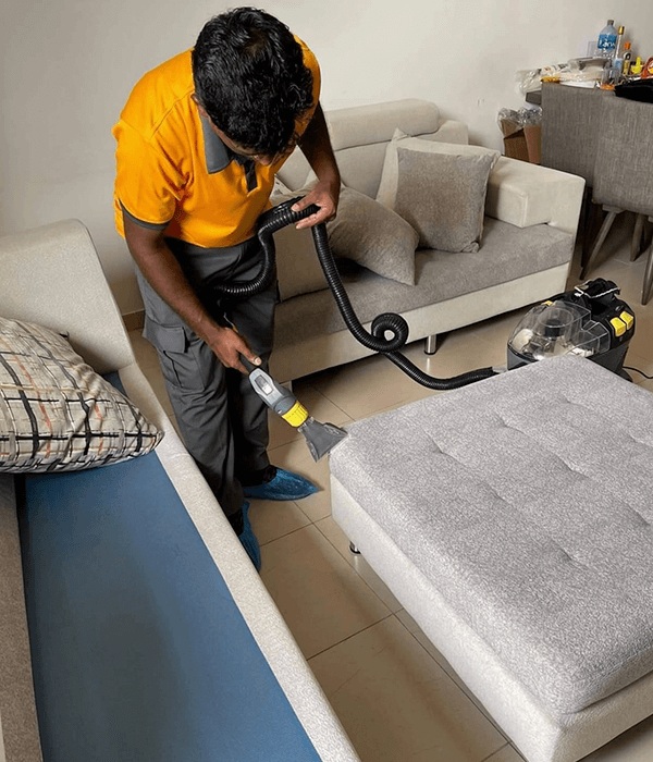 A person in a yellow shirt cleans a gray ottoman with a vacuum in a living room as part of maintenance service. Nearby are a beige sofa, pillows, and other household items.