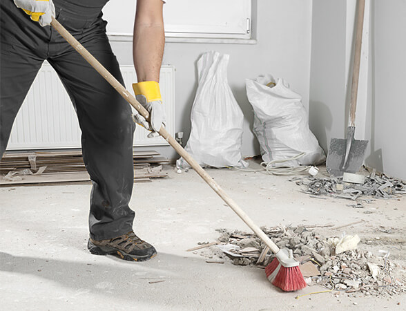 A person in work gloves sweeps debris with a broom in a room under renovation as part of specialized service. Nearby are piles of construction waste and tools, conveying a sense of labor.