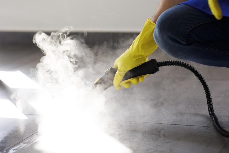 123 Person in yellow gloves using a steam cleaner on curtain cloth laid on the floor. Visible steam suggests thorough cleaning. Mood is focused and diligent.