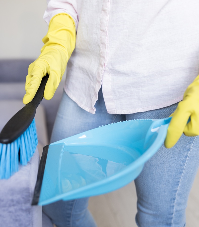 A person wearing yellow gloves cleans an upholstered sofa with a blue brush and dustpan, conveying a sense of tidiness and domestic chores.