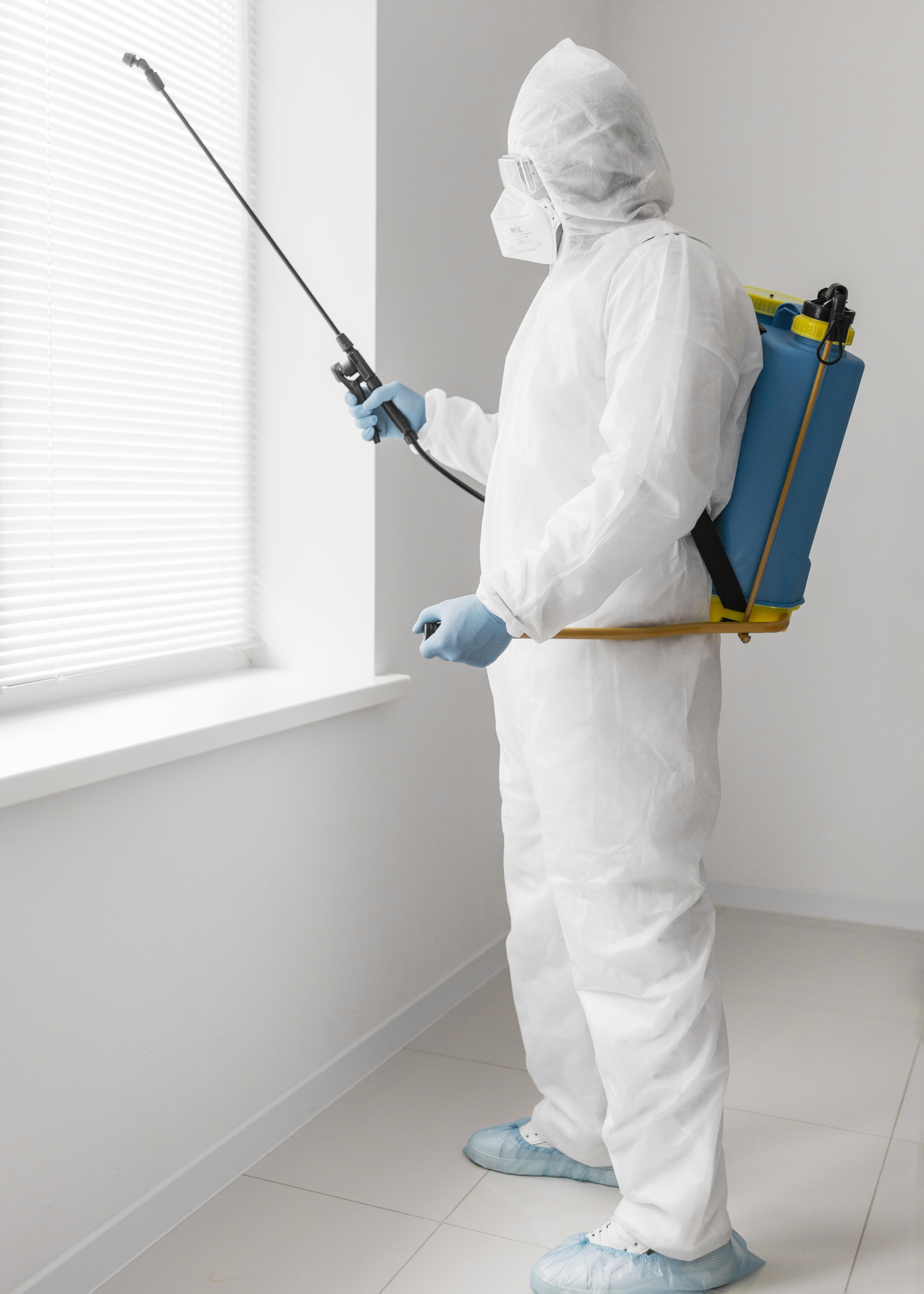 123 A person in protective suit and mask, holding a sprayer for pest control, standing by a window. They wear blue gloves and a backpack, suggesting sanitation work.