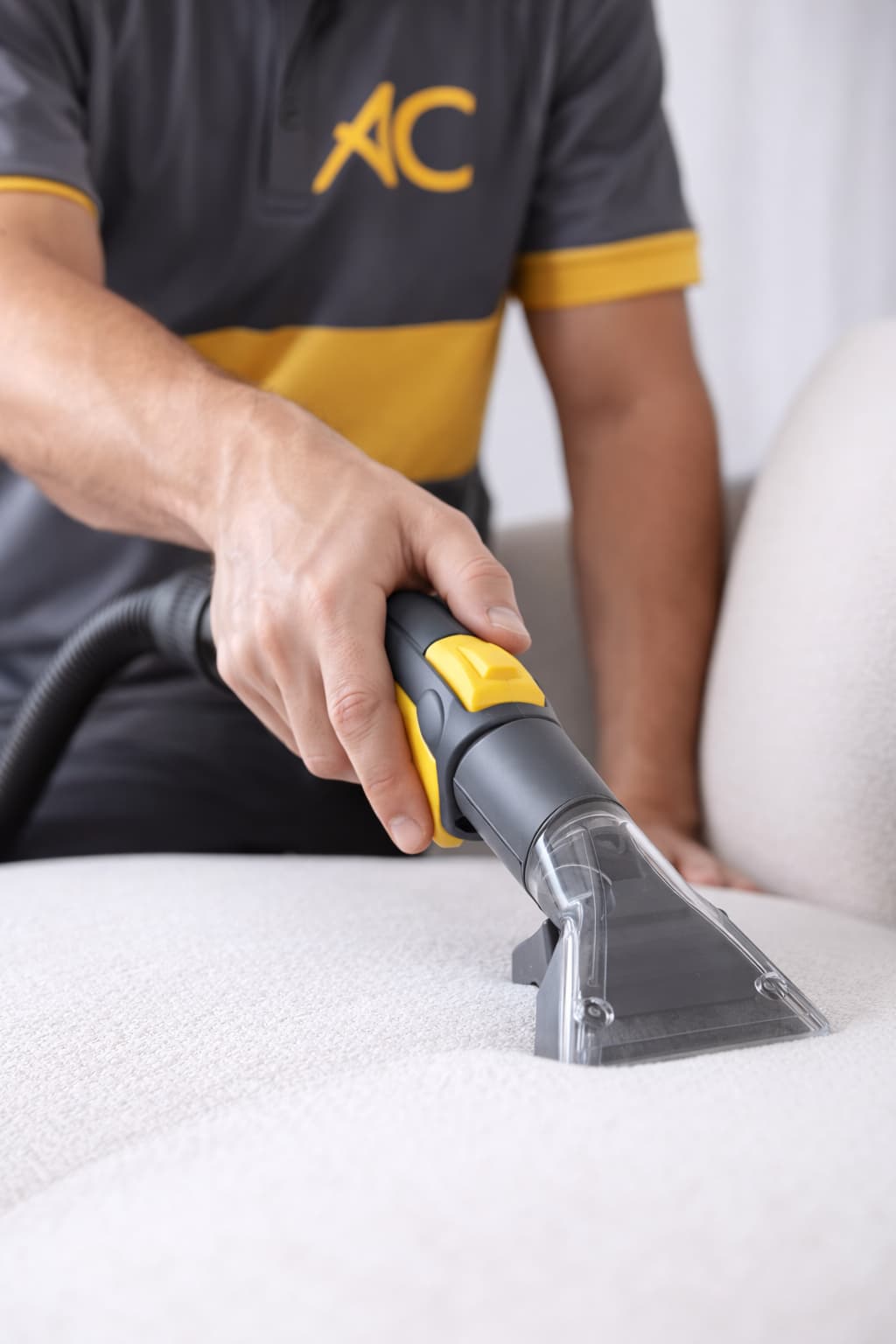 A close-up of a worker using a vacuum cleaner attachment to clean a white upholstered surface.