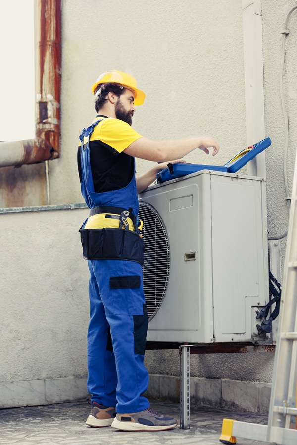 Worker in blue overalls and yellow hard hat repairing an outdoor air conditioning unit on a rooftop. The scene conveys focus and maintenance work.