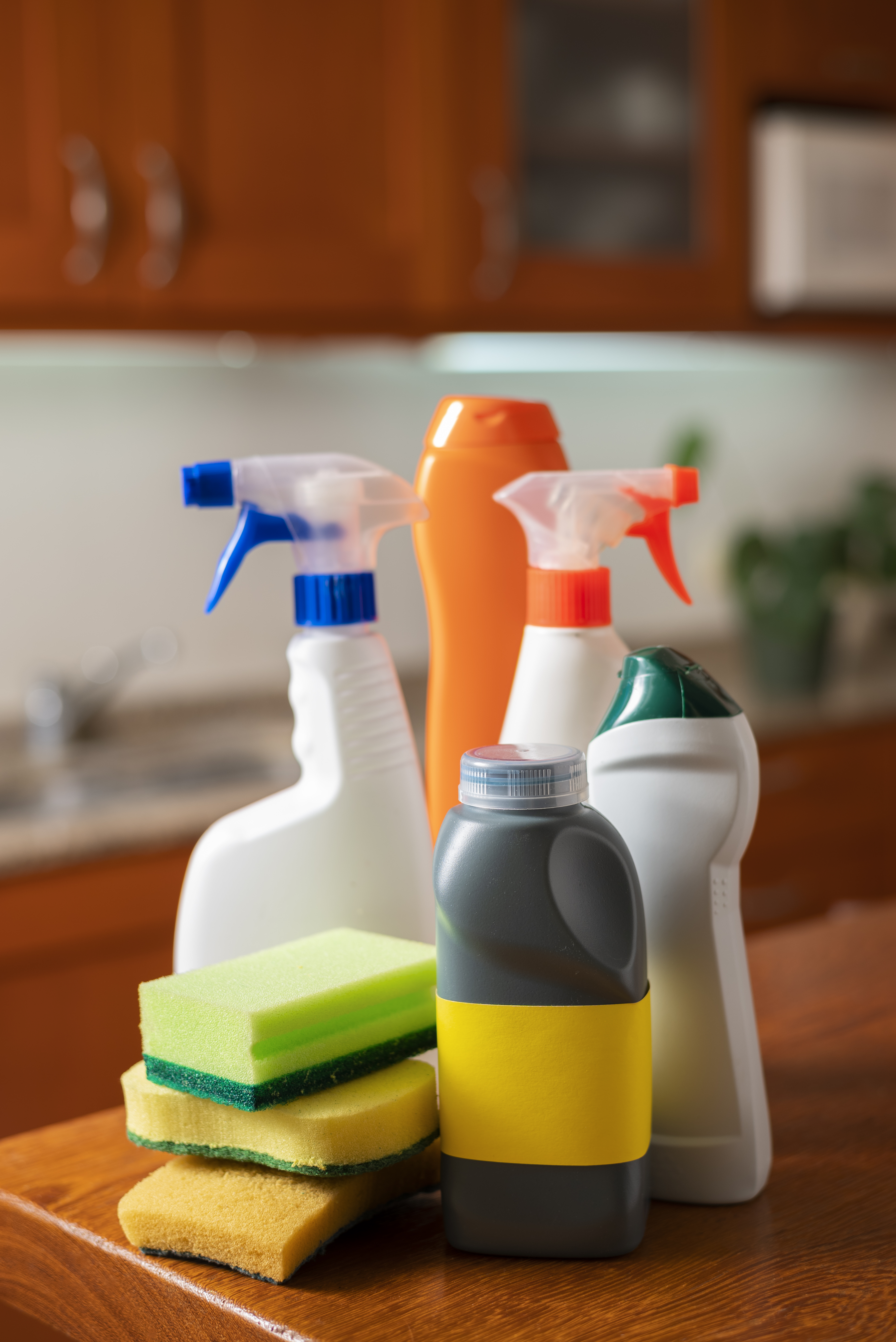 123 Cleaning supplies on a counter include spray bottles, sponges, and detergent, set against wooden cabinets and a blurred sink. Bright, organised setting.