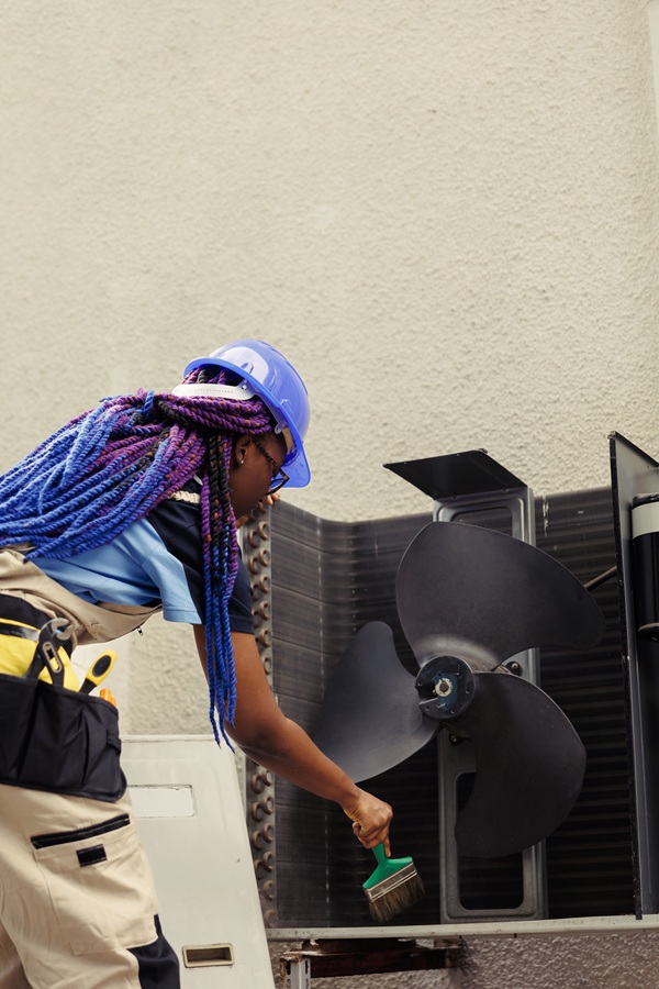 A technician with colorful braids and a blue hard hat cleans an air conditioning unit's fan with a brush. They carry tools in a belt, focused on the task.