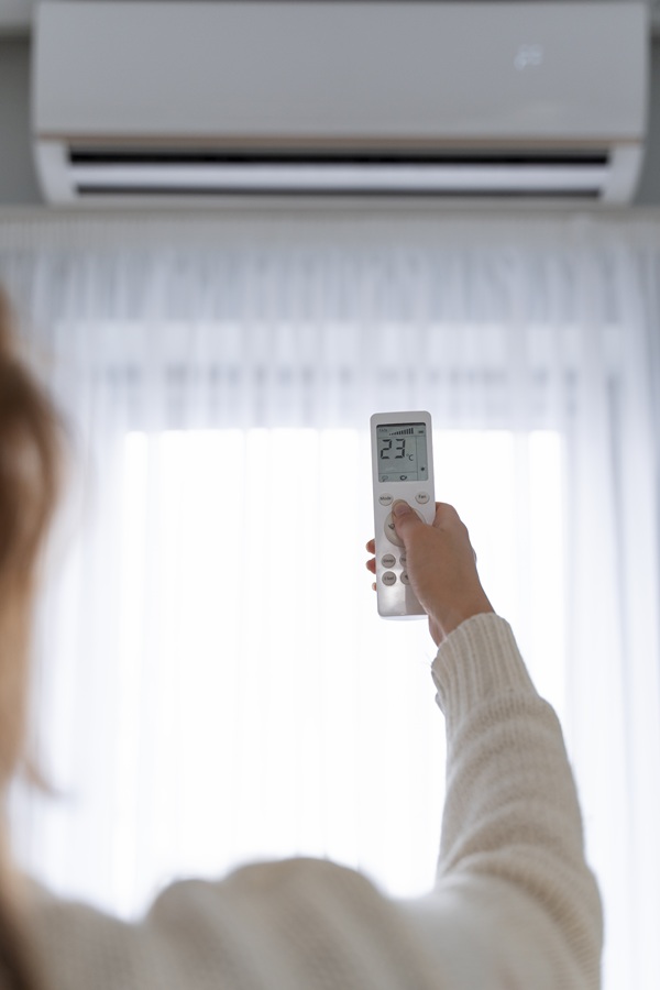 A person holds a remote control up towards an air conditioner mounted on the wall. The setting reads 23.1 degrees Celsius. Soft light filters through sheer curtains, creating a cozy atmosphere.