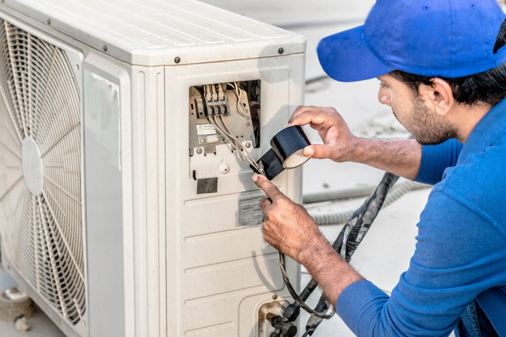 Man in blue cap and shirt repairing an outdoor air conditioning unit and its compressor, using black tape on exposed wires. The scene conveys focus and precision. 