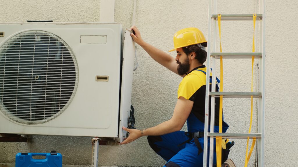 A technician in a yellow hard hat and blue overalls adjusts a wall-mounted air conditioning unit beside a ladder. The scene conveys professionalism and focus.