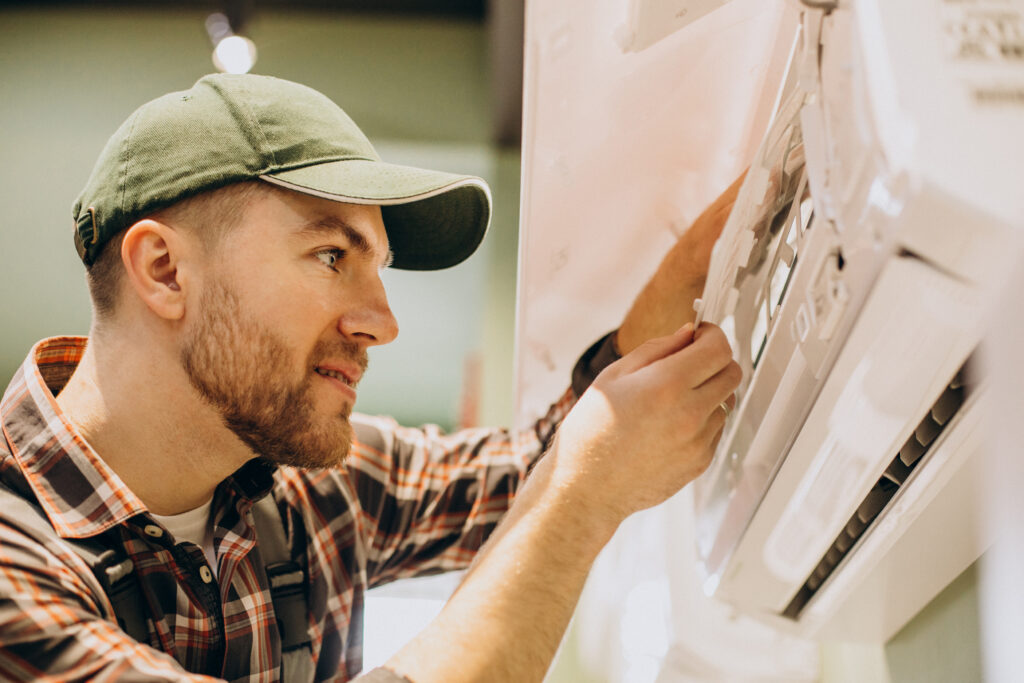 A man in a green cap and plaid shirt is focusing intently while fixing an air conditioner. The scene conveys a concentrated and professional atmosphere.