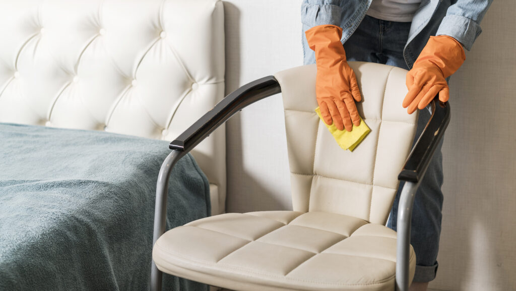 A person in orange gloves cleans a beige leather chair with a yellow cloth next to a bed with a blue blanket, conveying cleanliness and care