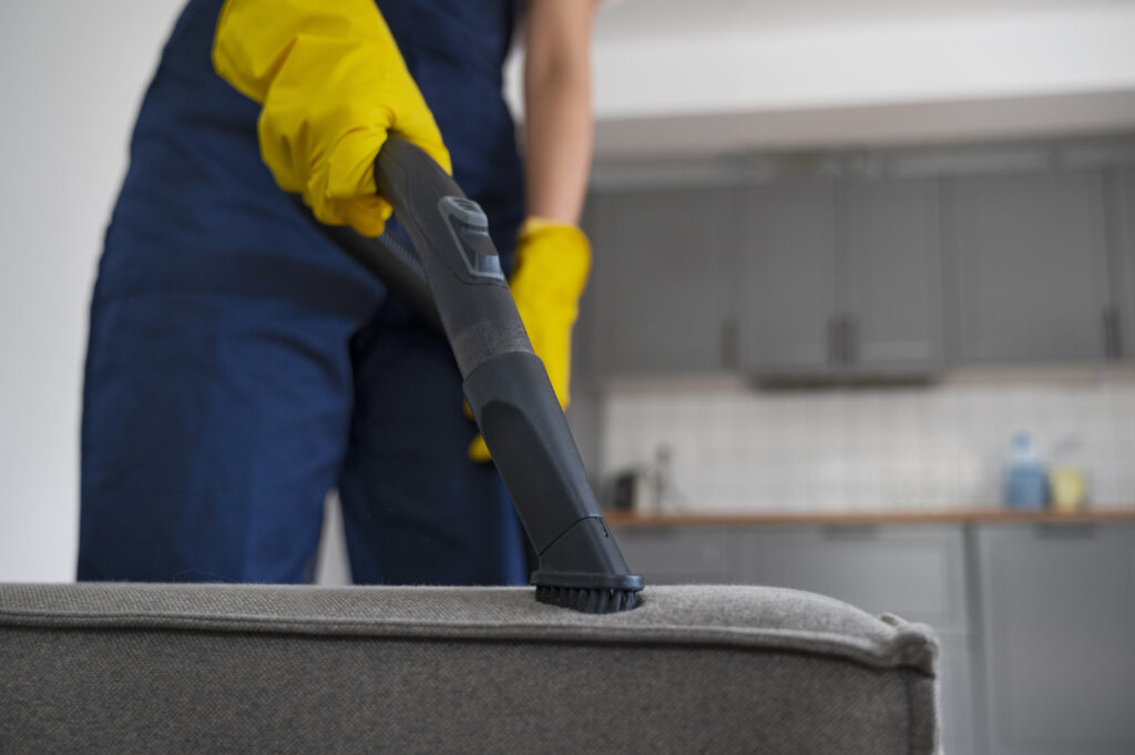 Person in blue overalls and yellow gloves vacuuming a gray sofa in a modern kitchen, conveying cleanliness and orderliness.