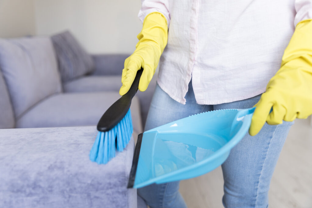 A person wearing yellow gloves cleans an upholstered sofa with a blue brush and dustpan, conveying a sense of tidiness and domestic chores.
