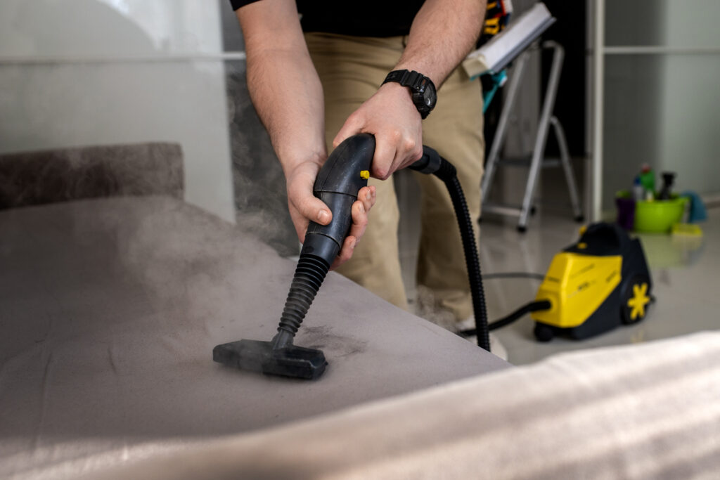 A person uses a steam cleaner on a couch, releasing visible steam. A yellow and black cleaning machine is in the background, adding an efficient, clean feel.