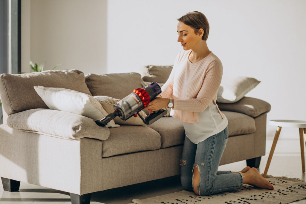 Person wearing yellow gloves and a blue apron cleans a beige sofa with a blue cloth and an orange spray bottle. Bright, clean, and tidy atmosphere.