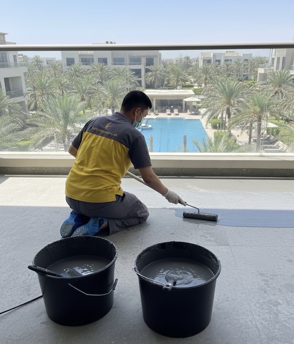 Technician brushes dark sealant over balcony tiles, completing vital waterproofing work by a sunny pool area.