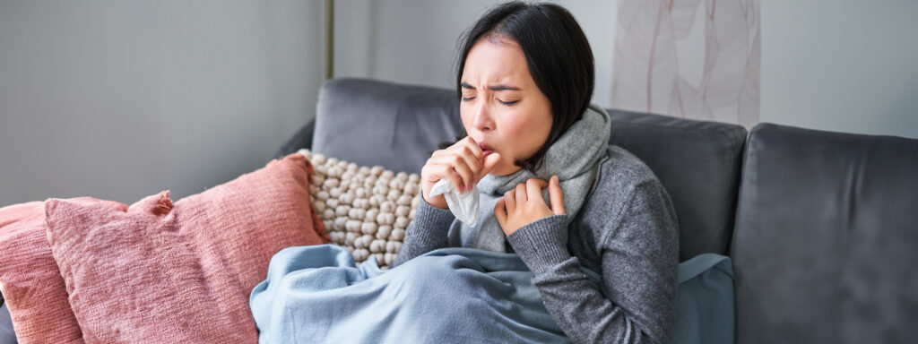 Asian woman coughing, feeling sick, sitting under blanket with scarf due to sore throat caused by faulty AC unit.